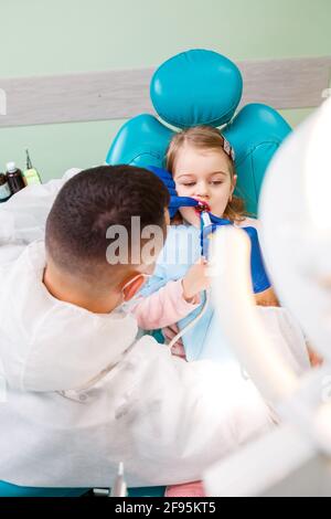 Un medico professionista, un dentista per bambini, tratta i denti di una bambina con strumenti. Studio dentistico per esame paziente. Il processo di ammaccatura Foto Stock