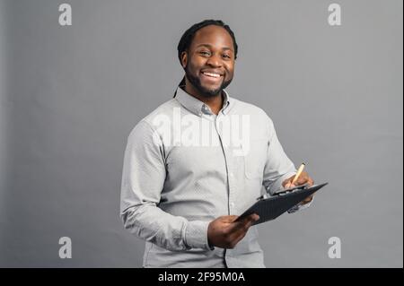 Giovane afroamericano allegro con dreadlock che tiene una cartella, prendendo appunti, sorridente uomo nero scrive giù con penna, isolato su sfondo grigio Foto Stock
