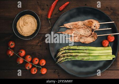 Piatto con spiedini di pollo alla griglia, asparagi verdi, salsa di arachidi, pomodori e peperoncino rosso su legno scuro. Vista dall'alto. Foto Stock