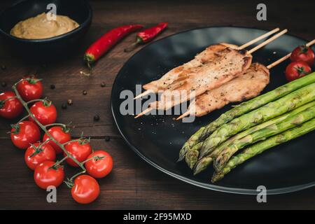 Piatto con spiedini di pollo alla griglia, asparagi verdi, salsa di arachidi, pomodori e peperoncino rosso su legno scuro. Foto Stock