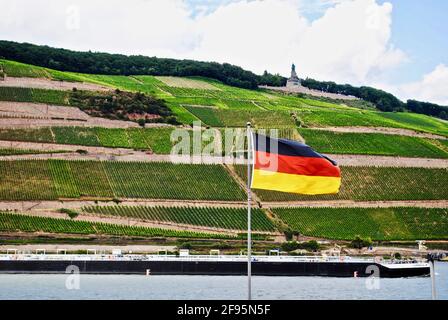 Bingen am Rhein, Germania: Fiume Reno con una barca, bandiera tedesca e vigneti. Niederwalddenkmal, statua di Germania, un monumento per la riunificazione tedesca. Foto Stock
