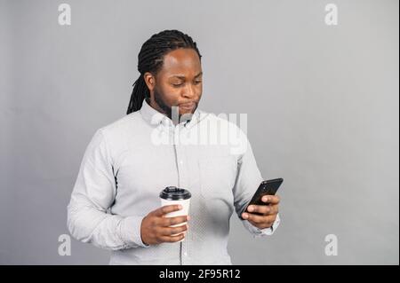 Uomo d'affari afro-americano serio, occupato e sicuro che indossa magliette e occhiali testando sullo smartphone, tiene una tazza di carta di caffè da asporto, dipendente di ufficio maschile isolato Foto Stock