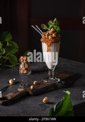 Ancora vita con delizioso frullato di grandi dimensioni in grande vetro con popcorn, caramello, menta e due cannucce di metallo in piedi su tavola di legno e su linguetta nera Foto Stock