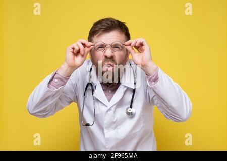 Il medico divertente con la vista povera tiene gli occhiali nelle mani e cerca di vedere qualcosa, con un'espressione sorpresa sul viso. Foto Stock