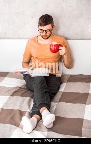 Il giovane legge un libro nel letto e beve il caffè. Foto verticale Foto Stock