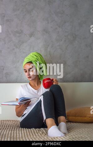 la giovane donna si siede su un letto con un libro e una tazza di caffè. Una donna con una maschera sul viso e un asciugamano sulla testa in abiti domestici. Foto Stock