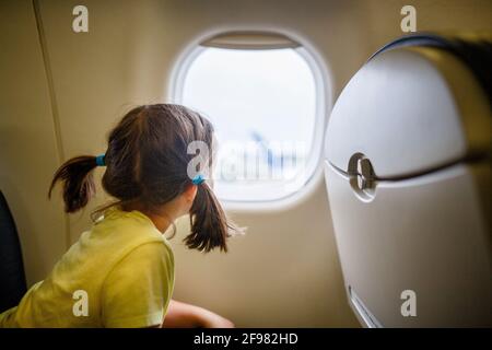 La bambina con i pigtail si siede sull'aereo fissando fuori la finestra su asfalto Foto Stock