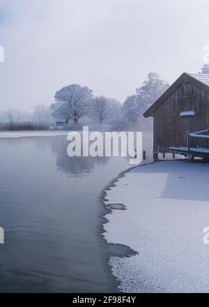 Una magica mattinata invernale alle boathouses di Schlehdorf am Kochelsee, Baviera, con brina, sole, nebbia e neve fresca caduta. Foto Stock