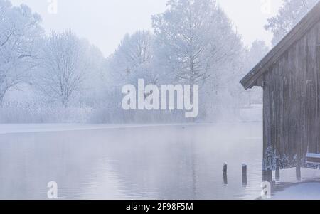 Una magica mattinata invernale alle boathouses di Schlehdorf am Kochelsee, Baviera, con brina, sole, nebbia e neve fresca caduta. Foto Stock