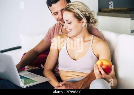 Stile di vita sano giovane coppia a casa godere di tecnologia e naturale cibo lavorando insieme su un computer portatile e mangiare una mela - persone in elazionship rfelice durante il blocco coronavirus Foto Stock