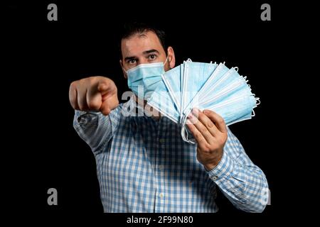 Uomo che indossa una maschera chirurgica per proteggersi dal coronavirus puntando alla fotocamera con una mano e tenendo più chirurgici maschere con l'altro Foto Stock