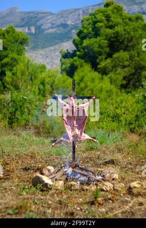 Agnello intero asado, barbecue su croce di ferro spiedo accanto al fuoco aperto in Altea la Vella, Alicante, Spagna Foto Stock