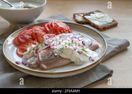 Filetti tradizionali di aringhe salate olandesi chiamati anche maatje con salsa alla crema, cipolle rosse, guarnitura di aneto e insalata di pomodoro servita su un piatto di legno Foto Stock