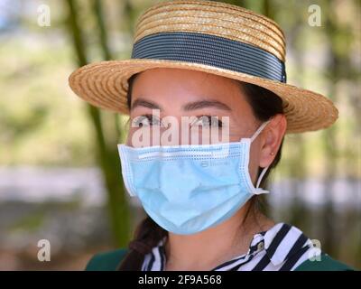 Bella giovane donna messicana indossa un elegante cappello di paglia Panama e si pone con una maschera chirurgica blu chiaro durante la pandemia del coronavirus globale. Foto Stock