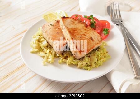 Bistecche di tonno su cremoso cavolo sabaudo con pomodori, fette di limone e guarnitura di prezzemolo su un piatto bianco e un tavolo di legno chiaro con tovagliolo e posate, Foto Stock