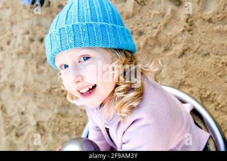 Il bambino si siede sul dispositivo di gioco e sorride alla macchina fotografica, ragazza bionda, serie di ritratti Foto Stock