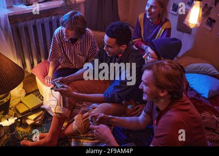 Gruppo di amici seduti sul pavimento in camera oscura utilizzo dei joystick durante la riproduzione di un videogioco in una festa Foto Stock