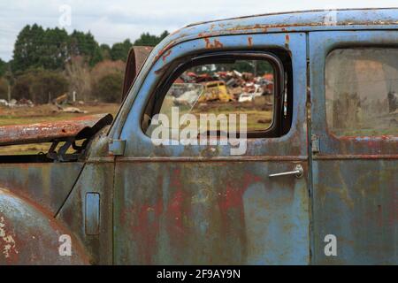 Una vecchia auto blu arrugginita nel cortile di un relitto, con altri veicoli rottamati visibili attraverso i finestrini rotti dell'auto Foto Stock