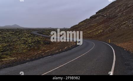 La prospettiva di una strada di campagna vuota e tortuosa (a forma di S) accanto al campo lavico coperto di muschio di pietre vulcaniche vicino a Grindavik, Reykjanes, Islanda. Foto Stock