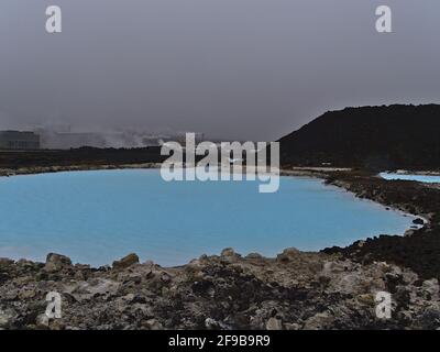 Piscina con acqua termale di colore blu tra le rocce laviche con la centrale elettrica di Svartsengi in background vicino alla Laguna Blu, Grindavik, Islanda. Foto Stock