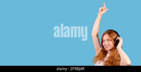 Banner, formato lungo su sfondo blu. Entusiasta donna caucasica che ascolta musica con le cuffie, ballando ritmicamente. Ragazza dai capelli rossi in posa Foto Stock