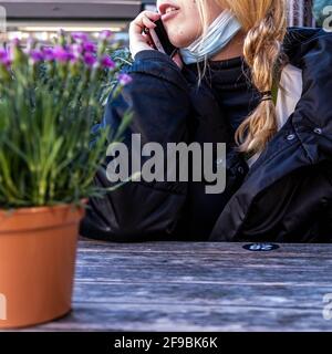 Epsom Surrey, Londra UK, aprile 17 2021, giovane donna seduta da sola ad UN tavolo usando UN telefono cellulare che indossa UNA maschera protettiva del viso Foto Stock