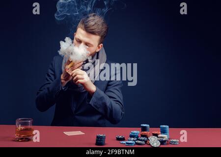 Elegante uomo con bearded in costume e sciarpa che gioca al casinò scuro, fumando sigaro, beve whiskey Foto Stock