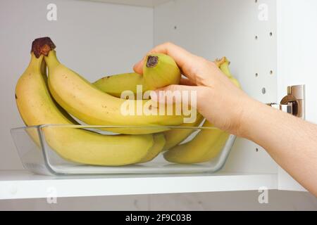 Una donna che prende un mazzo di banane da un vassoio di vetro che è in un armadio della cucina. Primo piano. Foto Stock