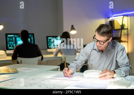 Concentrato maturo progettista automobilistico in occhiali seduta con modello di auto al tavolo e modifica dei layout in ufficio Foto Stock