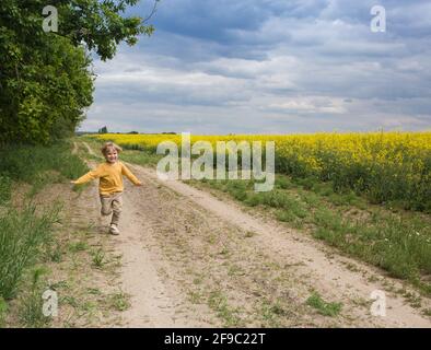 Sorridente simpatico bambino che corre spietatamente lungo una strada sterrata lungo un campo di colza giallo in primavera, felice infanzia, in avanti verso il futuro Foto Stock