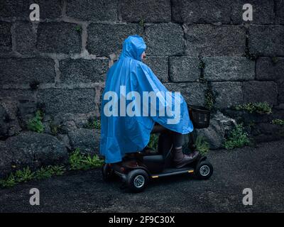 Uomo su uno scooter / veicolo portatile che indossa un impermeabile blu / Poncho / Cagoule / giacca, in profilo con sfondo Old Grey Stone Wall Foto Stock