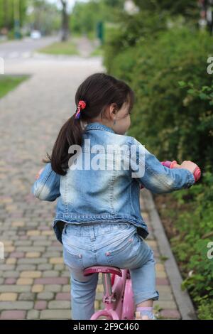 La bambina in un vestito in denim corre una bici rosa lungo un percorso in mattoni multicolore in una città di primavera Foto Stock