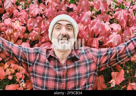 Ritratto di un uomo caucasico maturo vestito con una camicia controllata e un cappuccio di lana con un fondo di foglie autunnali. Foto Stock