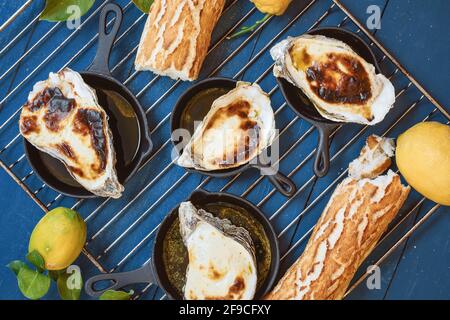 Ostriche cotte in casa in padelle di ghisa sul vecchio blu tavolo in legno decorato con limoni freschi con foglie di limone e. pane francese croccante Foto Stock