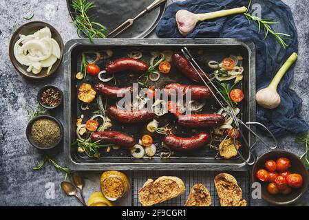 Vista dall'alto su deliziose salsicce alla griglia servite con cipolla, pomodori, aglio, pane ed erbe Foto Stock