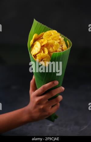 Chip di banana essiccata o cialcieri di banana, un cono di foglia di banana che tiene la mano riempito con cialciatori, fondo grigio testurizzato. Foto Stock