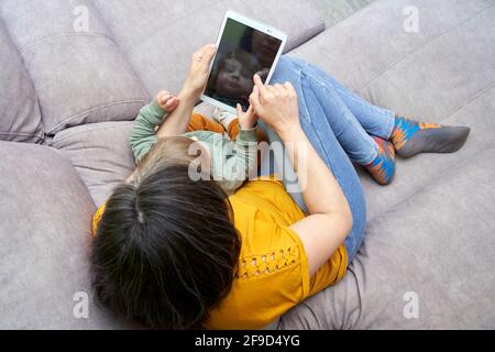 Una vista dall'alto della giovane donna con la figlia seduta sul divano a casa utilizzando un tablet. Concetto di tempo familiare, gadget, conciliazione, stile di vita. Foto Stock