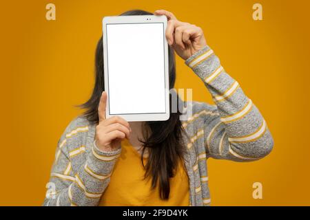 Una donna che ricopre il viso con un tablet pc su sfondo giallo dorato. Concetto di tecnologia, gadget. Foto Stock