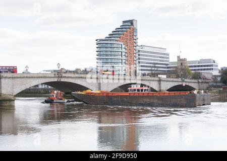 Una barca a rimorchiatore e una grande chiatta passano sotto il Putney Bridge con il blocco di appartamenti della Putney Wharf Tower sullo sfondo, Londra, SW15, Inghilterra, Regno Unito Foto Stock