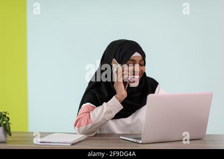 donna afro musulmana che indossa un hijab si siede sorridente in lei ufficio domestico e conversazione sullo smartphone Foto Stock