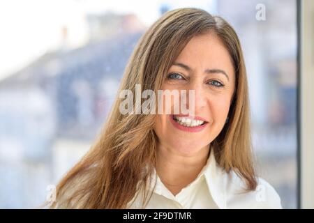 Donna di mezza età che guarda la fotocamera in modo silenzioso sorridi in un primo piano testa e spalle contro cui è stato girato una grande finestra Foto Stock