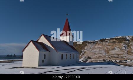 Splendida vista della piccola chiesa situata sopra il villaggio di Vík í Mýrdal sulla costa meridionale islandese con l'oceano atlantico e la formazione rocciosa di Reynisdrangar. Foto Stock