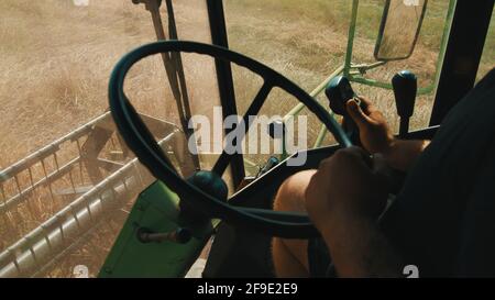 Vista interna di una mietitrebbia. Agricoltore maschio che manipola il volante in un campo agricolo. La barra falciante della mietitura può essere vista tagliare i raccolti durante il giorno. Foto Stock