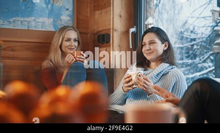 Vacanza invernale. Le amiche si siedono vicino alla finestra in un'accogliente casa di legno e bevono bevande calde. Foto di alta qualità Foto Stock
