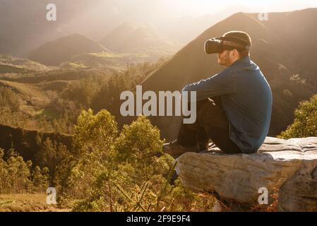 Vista laterale del viaggiatore maschile in occhiali VR con cui interagisce la realtà virtuale mentre si siede su una collina su un terreno montuoso a. tramonto Foto Stock