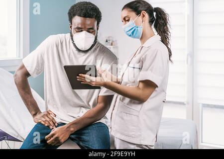 Giovane dottoressa in uniforme medica e stetoscopio che indossa la faccia Maschera che parla e mostra il risultato su tavoletta ad afroamericano paziente uomo durante Foto Stock