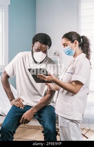 Giovane dottoressa in uniforme medica e stetoscopio che indossa la faccia Maschera che parla e mostra il risultato su tavoletta ad afroamericano paziente uomo durante Foto Stock