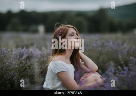 Vista laterale di una donna delicata con fiori in pelo seduto in fiore campo di lavanda e godendo la natura con gli occhi chiusi Foto Stock