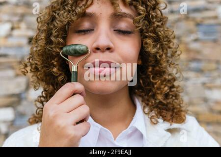 Sognante donna afroamericana usando il rullo di giada e facendo il viso massaggio in piedi con gli occhi chiusi nel parco Foto Stock