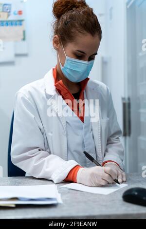 Medico femminile serio in maschera e uniforme seduto a tavola in sala medica e la prescrizione di scrittura su carta Foto Stock